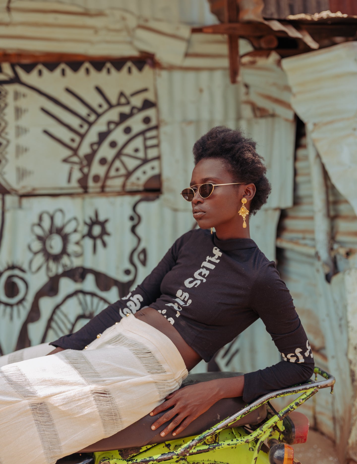Model wearing LilaBare’s black Gujarati Script Crop Top, featuring long sleeves and a high neckline with “Made for the Mindful” printed in Gujarati script, paired with cream handloom pants while seated on a motorbike in front of a corrugated metal wall with graffiti.