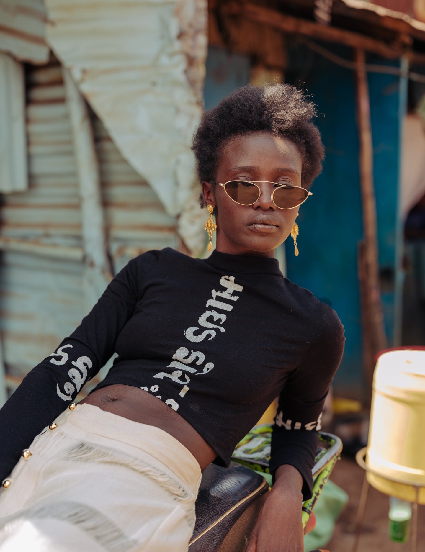 Model wearing LilaBare’s black Gujarati Script Crop Top, featuring long sleeves and a high neckline with “Made for the Mindful” printed in Gujarati script, paired with cream handloom pants while seated on a motorbike in front of a corrugated metal wall with graffiti.