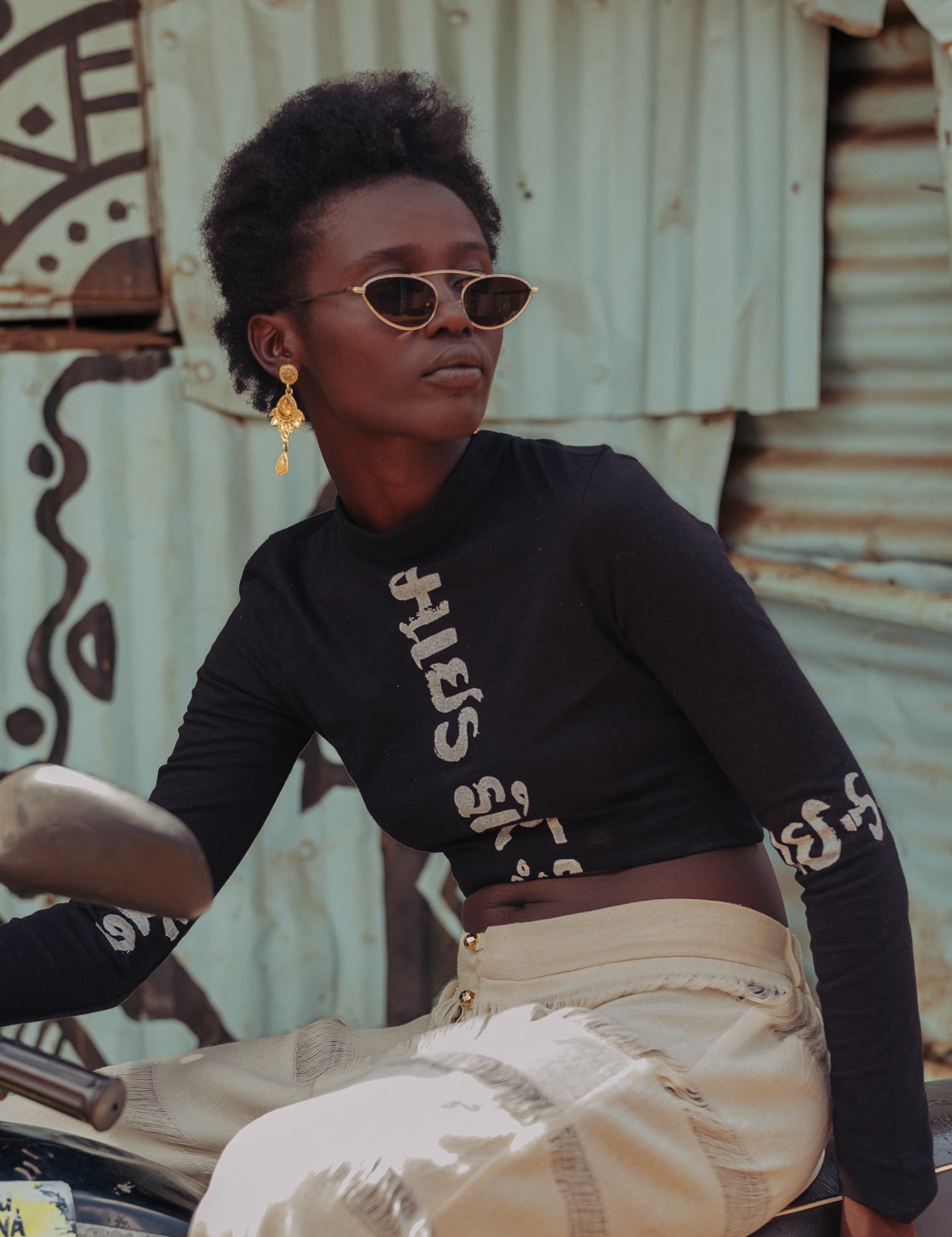 Model wearing LilaBare’s black Gujarati Script Crop Top, featuring long sleeves and a high neckline with “Made for the Mindful” printed in Gujarati script, paired with cream handloom pants while seated on a motorbike in front of a corrugated metal wall with graffiti.