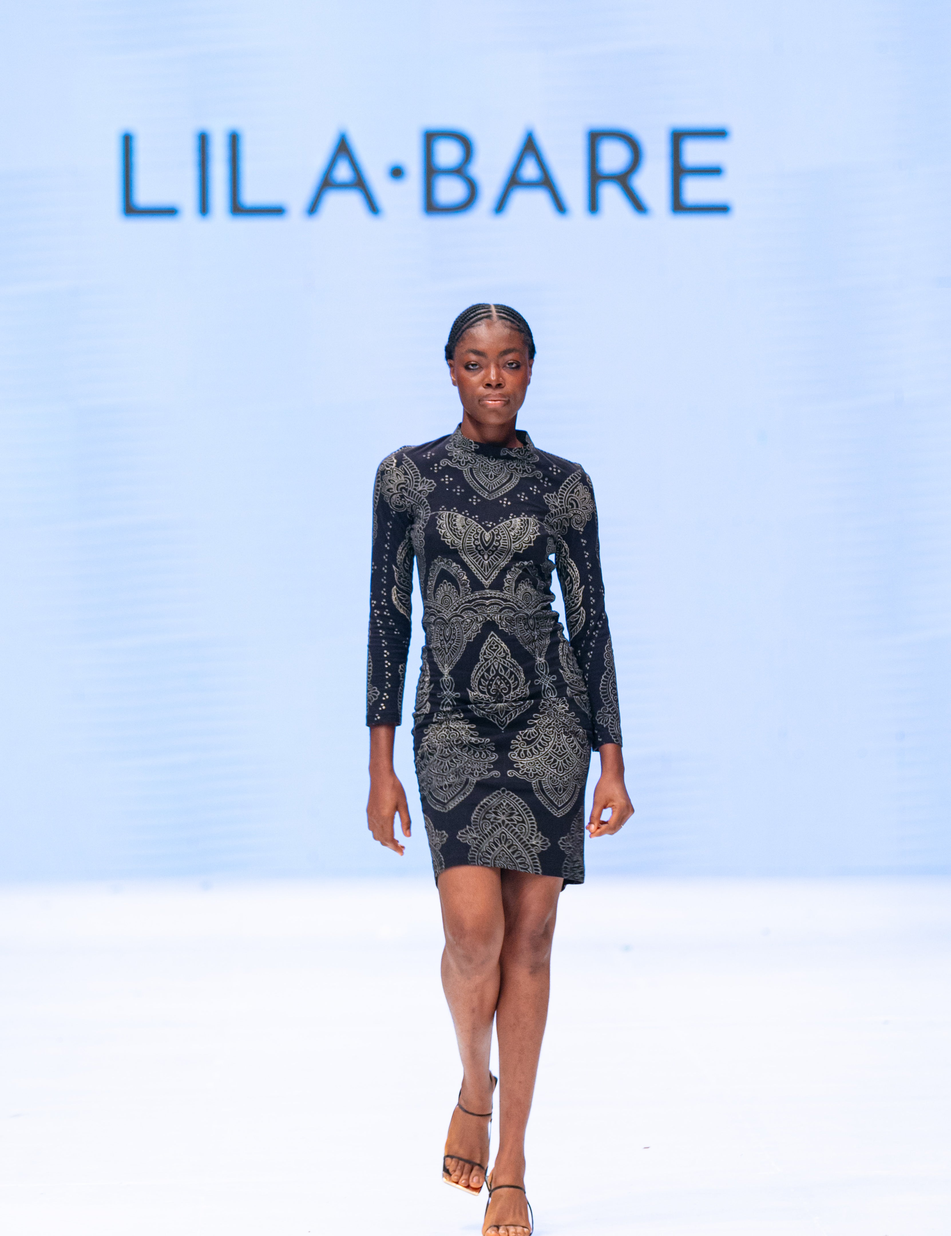 Model walking runway in black form-fitting Mehendi Cocktail Dress with long sleeves, featuring digitally engineered laser-engraved blockprint design in natural cotton jersey by LilaBare.