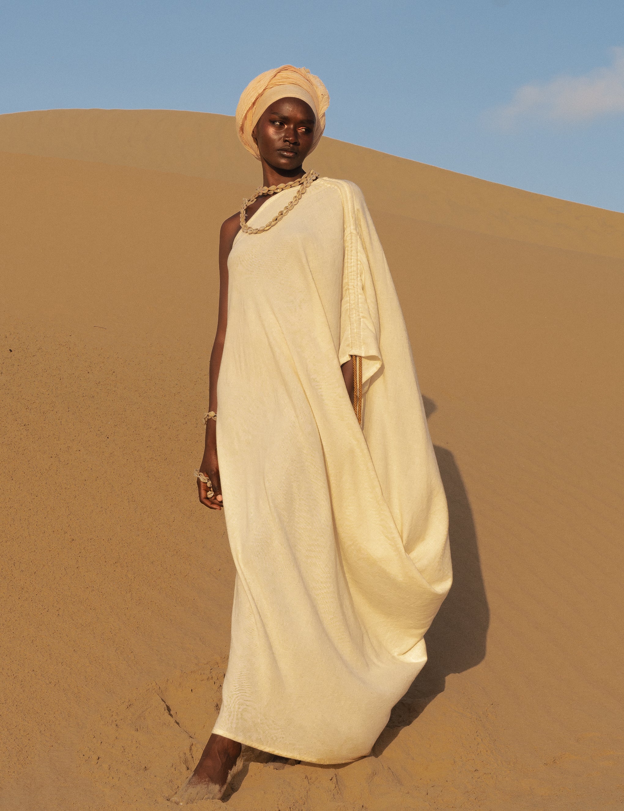 Model wearing LilaBare’s One Shoulder Sari Dress in undyed handloom cotton, walking barefoot in golden desert sand at sunrise.