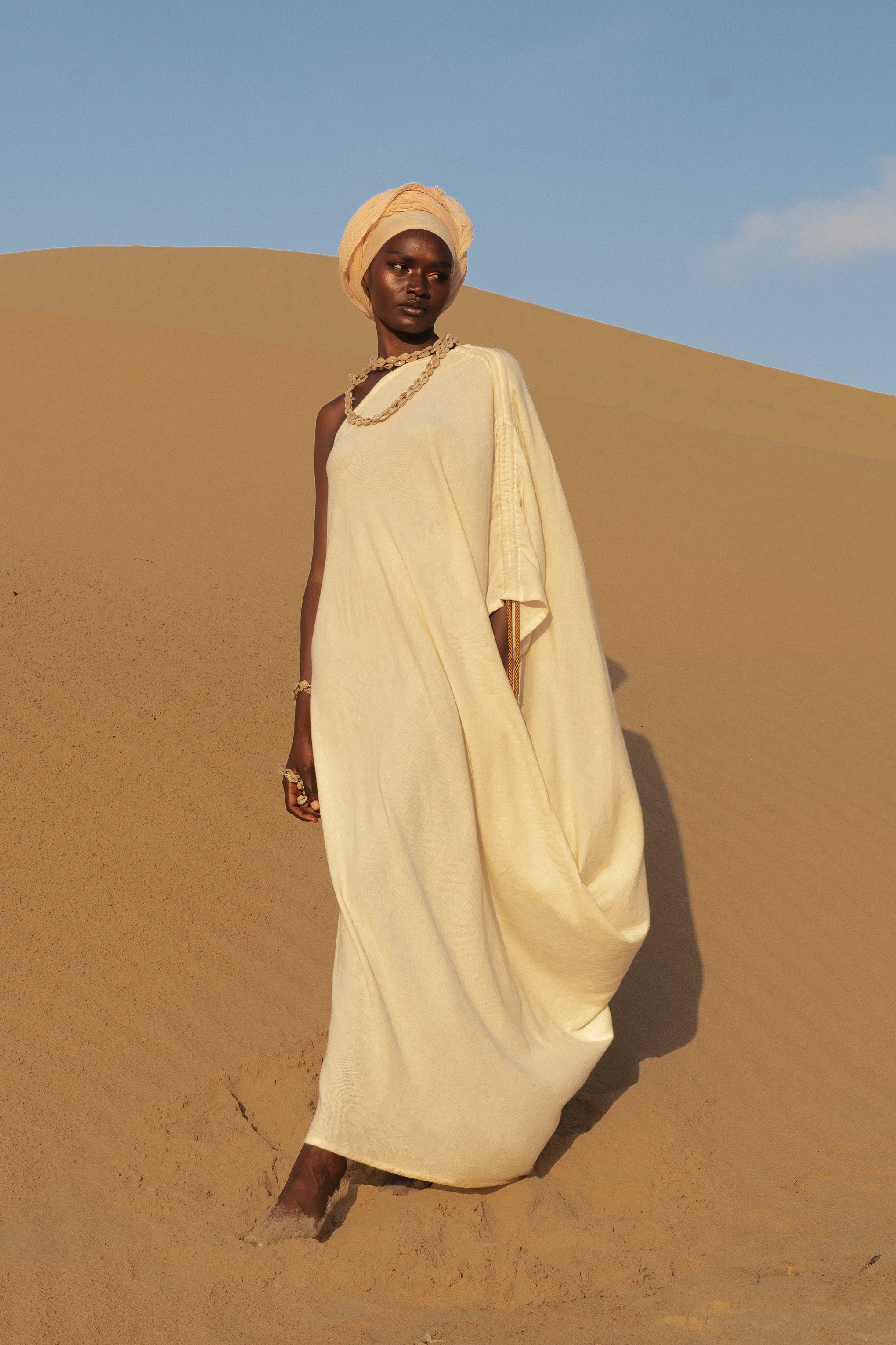 Model wearing LilaBare’s One Shoulder Sari Dress in undyed handloom cotton, walking barefoot in golden desert sand at sunrise.