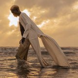 Model wears the Shakti Shirt and wide-leg pants in a ocean setting, showcasing relaxed silhouette and gender-fluid style.
